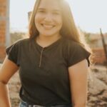 A cheerful young woman stands outdoors at sunset, backlit by warm sunlight.